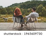 Back view of young couple sitting with the dog in front of their beautiful wooden country house. Elegant couple sitting on chairs holding hands and enjoying beautiful mountain view. Stay home concept 