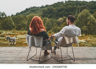 Back View Of Young Couple Sitting With The Dog In Front Of Their Beautiful Wooden Country House. Elegant Couple Sitting On Chairs Holding Hands And Enjoying Beautiful Mountain View. Stay Home Concept 