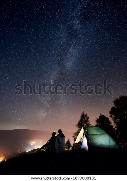 Back view of young couple tourists having a rest at\
bonfire near illuminated tent under amazing night sky full of stars\
and Milky way. On background beautiful starry sky, mountains and\
luminous city