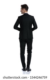 back view of young elegant in tuxedo looking to side and walking isolated on white background, full body