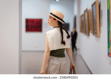 Back view of a young elegantly dressed woman in a straw hat admiring the exhibition of paintings in the gallery. Contemporary art at the Art Museum.