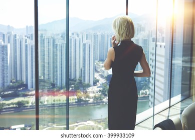 Back view of young entrepreneur is calling via cell telephone, while is standing in her private office against window with view of developed business district with high skyscrapers.Copy space for text