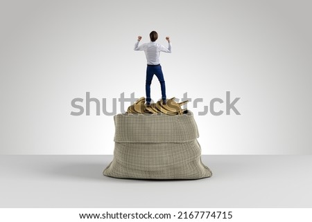 Back view of young european businessman celebrating success on huge sack filled with golden coins on light background. Wealth, treasure and boss concept