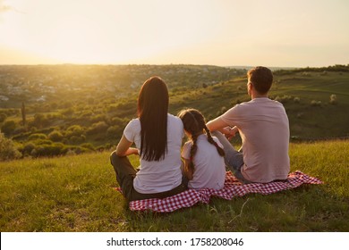 Back view of young family with child sitting on picnic blanket and looking at beautiful sunset in mountains, copy space
