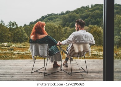 Back View Of Young Family Travelers Holding Hands, Sitting On The Terrace. Happy Man And Woman Sitting And Relaxing On Wooden Porch Of Modern House And Enjoying Beautiful View On Forest In Mountains