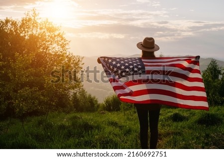 Back view of young female 20s old years in hat holding national American flag on background summer mountains. Holiday travel and romantic amazing sunset. America independence day, July 4th concept