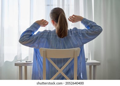 Back View Of Young Female Sitting At Desk And Stretching. Remote Work Challenges And Negative Aspetcs. Working At Home Problems. Downsides Of Hustle Culture. Self-distancing Workspace. Neutral Light.