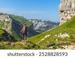 Back view of young female traveler with trekking sticks hiking in Alps. Woman tourist walking alone on mountain path overlooking green alpine valley. Summer mountain hiking and active people concept.