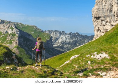 Back view of young female traveler with trekking sticks hiking in Alps. Woman tourist walking alone on mountain path overlooking green alpine valley. Summer mountain hiking and active people concept. - Powered by Shutterstock