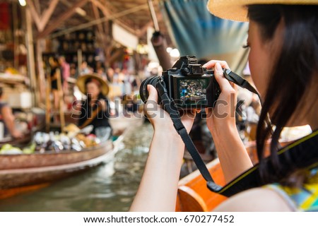 back view of young girl traveler using camera taking photo with local vendor when she taking on boat visiting Damnoen Saduak floating market of Thailand.