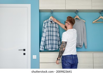 Back view of young guy in casual clothes taking checkered shirt from wardrobe while dressing in morning at home - Powered by Shutterstock