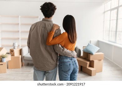 Back view of young guy with his girlfriend hugging each other in living room of new house on moving day. Millennial Asian couple embracing in their owned apartment. Relocation concept