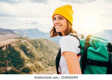 Back View Young Happy Smiling Woman Backpacker With Brunette Hair Relaxation On Top Rocky Mountains, With Backpack, Yellow Hat. Leisure After Walking Valley. Adventure, Travel, Holiday Concept.