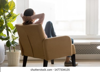 Back view of young happy woman sitting in comfortable armchair, facing shiny window, crossing hands behind head, having rest after housework, resting on weekend, lazy day concept.