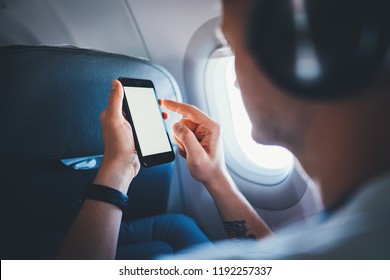 Back view of young hipster guy sitting in aircraft using modern cellphone and listening to music by headphones, professional businessman working on smartphone device during flight, business travel 
