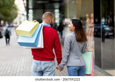 Back view of young international couple walking hand in hand near big mall, holding shopper bags outdoors. Millennial multiracial family window shopping, enjoying their walk on city street - Powered by Shutterstock