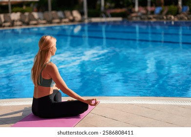 Back view of a young long haired blonde in sportswear sitting on an outdoor yoga mat by the pool. Yoga time. Relaxed woman meditating in lotus position, yoga practicing.