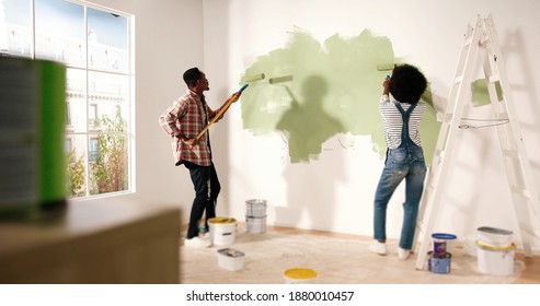 Back View Of Young Lovely African American Family Married Couple Doing Renovating Works In House. Wife And Husband Painting Walls Together Using Roller Brushes Redecorating And Redesigning Home