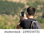 Back view of a young man with backpack taking photos of the mountain view with his smartphone, close up, male hiker photographing nature with his cellphone, guy in the forest taking pictures