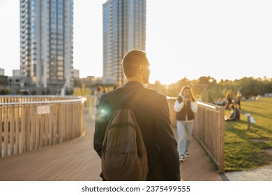Back view of young man with backpack standing on wooden bridge and looking at city