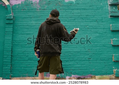 Back view of young man in black hoodie standing in front of green brick wall of building before spraying paint and creating artowrk