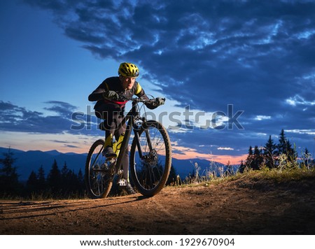 Back view of young man cycling bicycle under beautiful night sky. Male bicyclist in safety helmet riding on hillside road under blue cloudy sky in the evening. Concept of sport and active leisure.