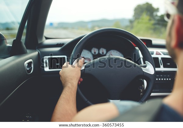 Back view of a young man driving his car during\
summer trip, hipster guy in sunglasses enjoying a road trip during\
vacation time, male driver sitting at steering wheel with copy\
space for your brand
