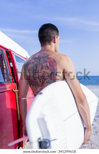 back view of a young man is holding a surf board\
next to a classic van on a surf session on the beach - focus on the\
girl tattoo face