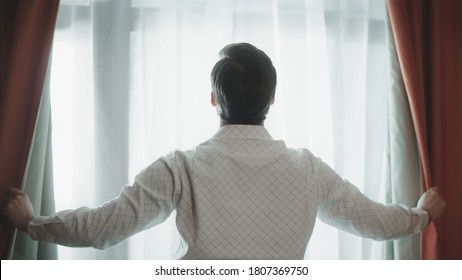 Back View Of Young Man Opening Curtains At Hotel Room. Close Up Of Handsome Business Man Standing By Window. Successful Businessman Opening Curtains At Modern Apartment