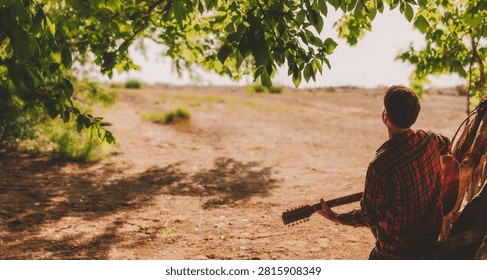 Back view of young man playing acoustic guitar while looking out at river from under forest trees. Concept of creative inspiration, freedom, and authentic lifestyle at riverside camp.