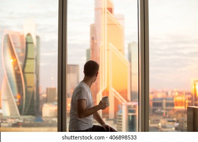 Back View Of Young Man Sitting On Window With Coffee Cup, Looking At Dawn City Scenery After Early Wakeup. Handsome Casual Guy Relaxing In Penthouse After Good Night Sleep And Watching Sunrise