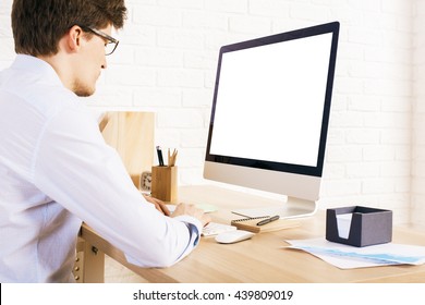 Back View Of Young Man Sitting At Office Desk Using Computer With Blank White Monitor. Mock Up