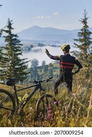 Back view of young man standing on grassy hill near bicycle and pointing finger at mountains. Bicyclist gesturing towards foggy hills. Concept of sport, bicycling and active leisure.