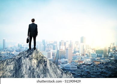 Back view of young man in suit and with briefcase in hand standing on mountain top. City background with copy space. Leadership concept - Powered by Shutterstock