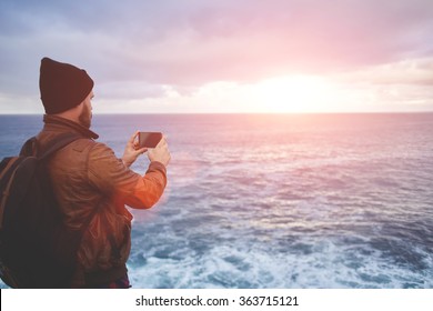 Back View Of A Young Man Tourist Taking Photo With Cell Telephone Digital Camera While Standing In Front Sea With Waves, Hipster Guy With Trendy Look Shoots Video With Ocean Landscape On Mobile Phone 