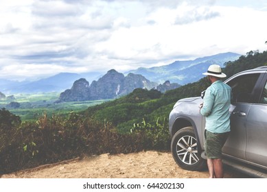 Back View Of A Young Man Wanderer Is Enjoying Amazing Jungle View, While Is Standing Near His Luxury Suv Car. Male Traveler Is Looking Away On The Beautiful Asian Landscape And High Mountain