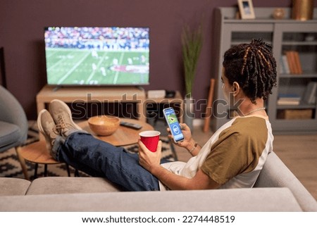 Back view at young man watching football match at home and relaxing on couch with online betting app