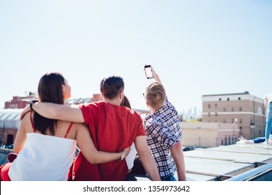 Back View Of Young Millennial People Hugging And Taking Selfie While Sitting Outdoors Against Cloudless Blue Sky On City Street Together. Group Of Friends Photographing Themselves On Smartphone Camera