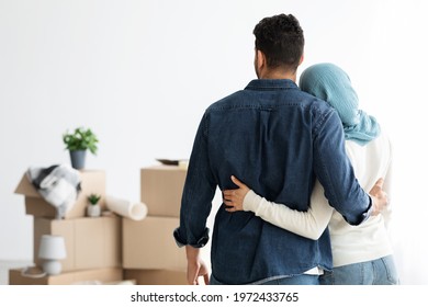 Back View Of Young Muslim Family Husband And Wife Cuddling While Moving To New House, Copy Space. Arab Man And Lady In Hijab Standing In Empty White Room, Looking At Paper Boxes And Hugging