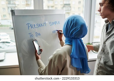 Back view of young Muslim female teacher explaining multi-ethnic guy Present Simple tense while wiriting down examples on whiteboard