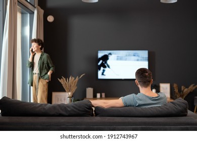Vista posterior de un joven reposado sentado frente al televisor y viendo noticias deportivas o transmitiendo mientras su esposa habla por teléfono