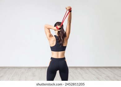 Back view of young sporty woman wearing black tank top and leggings, doing exercise for triceps with latex resistance band. Full length indoor shot against white wall. - Powered by Shutterstock