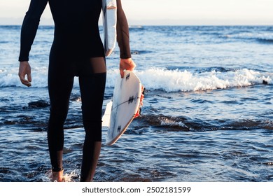 Back view of a young surfer in a wetsuit comes out of the water holding a broken surfboard. Horizon over the water and sunset light - Powered by Shutterstock