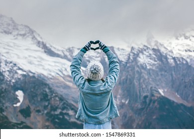 Vista traseira da jovem viajante em luvas e chapéu em pé sobre picos de montanhas nevadas e fazendo forma de coração de amor pelas mãos. Cena de viagem de inverno, conceito de desejo de viajar.