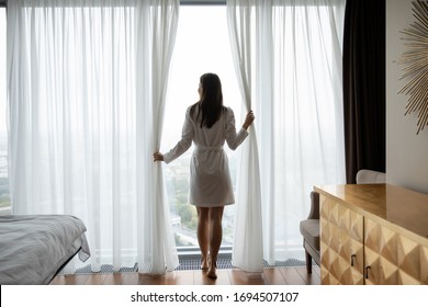 Back view of young woman in bathrobe wake up in modern bedroom open curtains enjoy good morning, millennial female awake at home or hotel feel positive optimistic welcome new sunny day