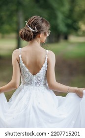 Back view of young woman with beautifully coiffured dark hair decorated with white floral hair clip holding edges of white dress, standing outside in park in summer. Soft focus, vertical. Wedding. - Shutterstock ID 2207715201