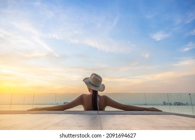 Back view of young woman in bikini with straw hat on the sun-tanned slim, shapely body with her arms spread to the side, relaxing in swimming pool on the roof top of hotel, enjoy cityscape at sunset.