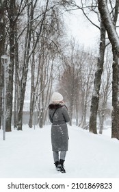 Back View Of Young Woman In Coat And Knitted White Hat Enjoying Walk In Winter Park, Outdoors. Rear View Of Lonely Girl Walking Along Snowy Alley. Vertical Image.