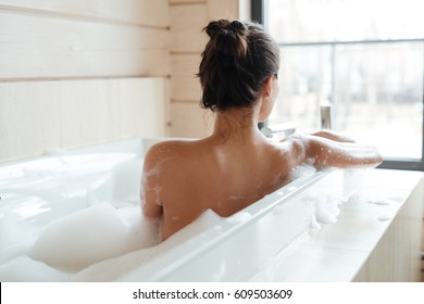 Back view of a young woman having bubble bath and looking at window