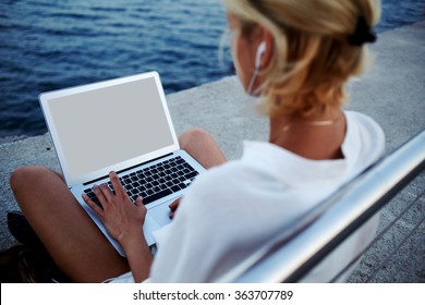 Back View Of Young Woman In Headphones Watching Movie On Laptop Computer While Sitting On Pier Near Sea, Female Tourist Using Net-book With Blank Copy Space Screen For Your Text Message Or Information